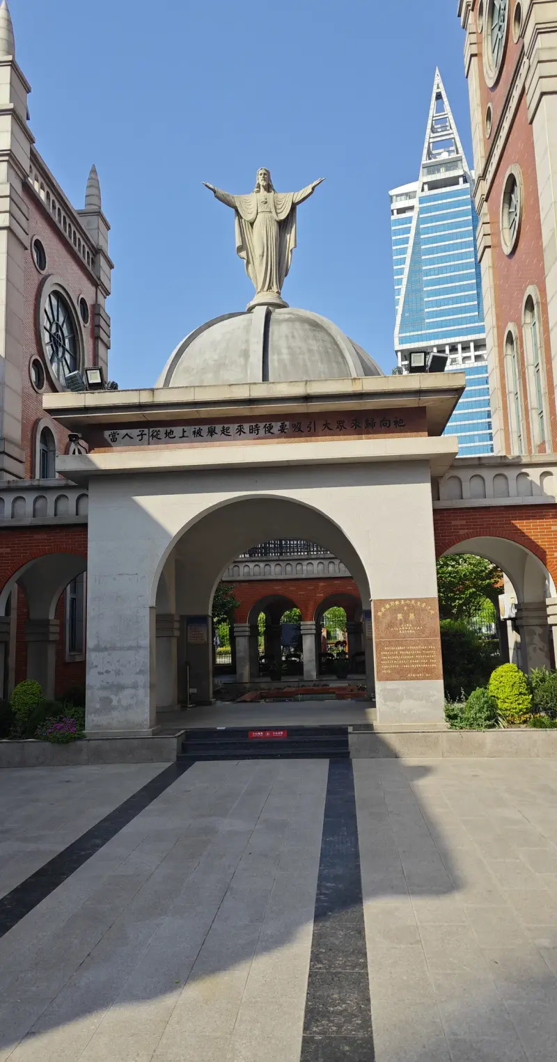 A pale stone gateway with a round arch and a dome carrying a statue with outstretched arms, with a brick tower behind.