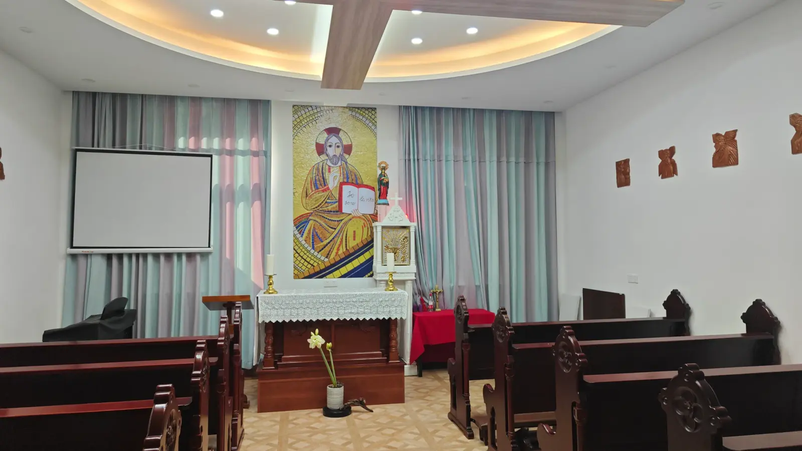 A small chapel with a lace-covered altar, a gold mosaic of a seated Christ, curtains and dark wooden pews.