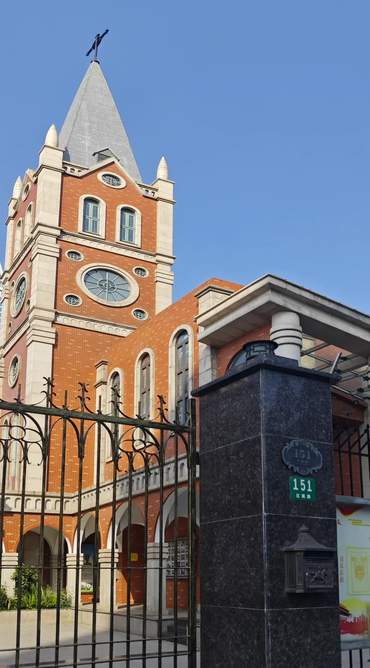A tall orange brick church tower with a clock and grey spire against a blue sky, with a dark granite gate post in the foreground.