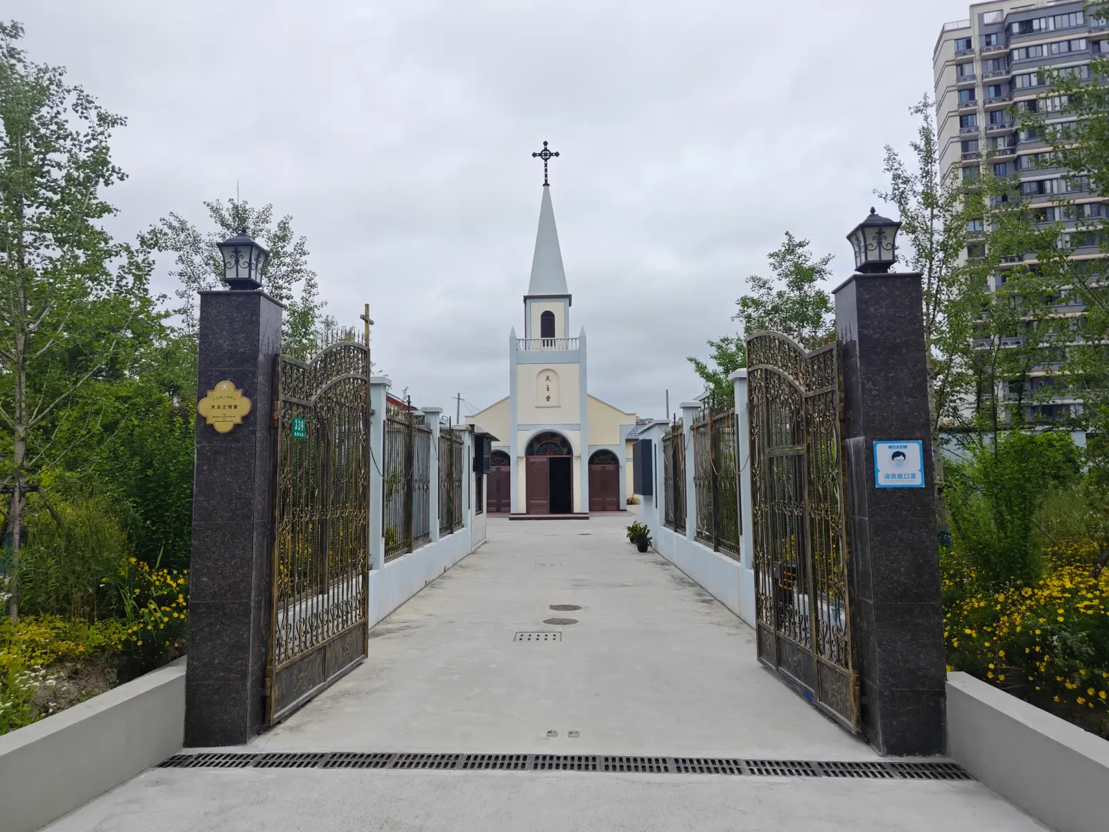A small white church with a tall spire and cross at the end of a concrete path between open iron gates and dark gate posts.