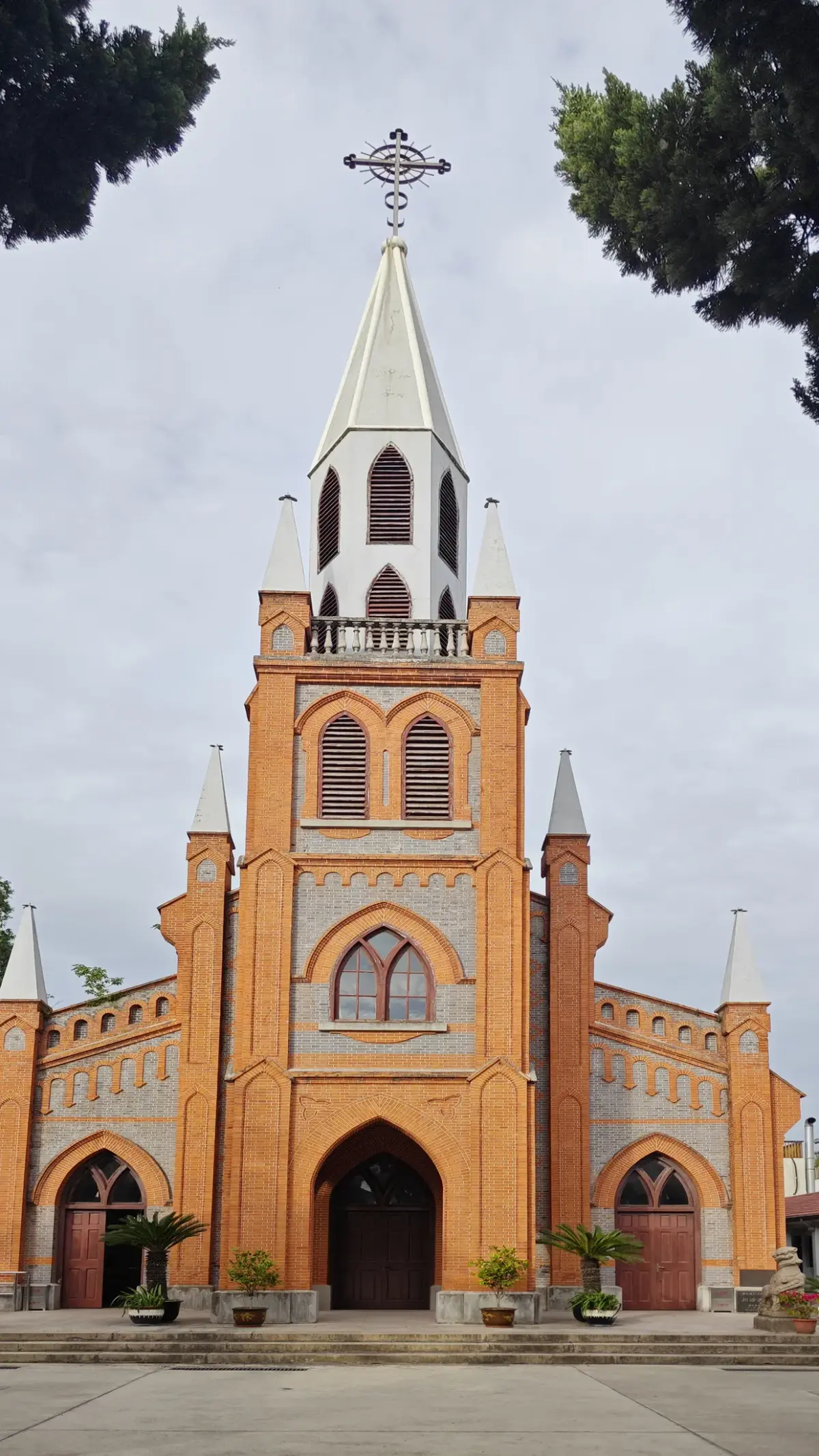An orange-brick church front with a central spire, pointed windows and three arched doors, framed by dark pine branches.