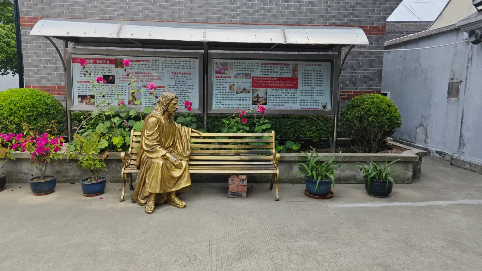 A gilded bronze statue of a robed seated figure on a park bench beside flowering shrubs and a covered notice board.