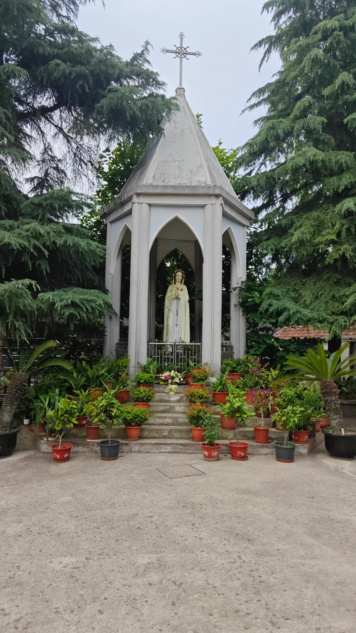 A grey pointed open shrine with a cross housing a white statue, among potted plants and pine trees.