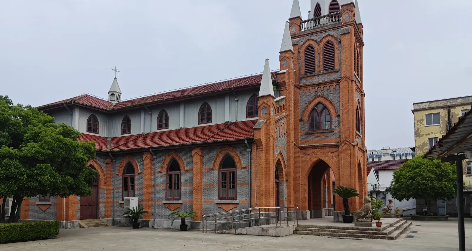 A long orange and grey brick church with pointed windows, a red tile roof and a tall entrance tower, seen across a paved courtyard.