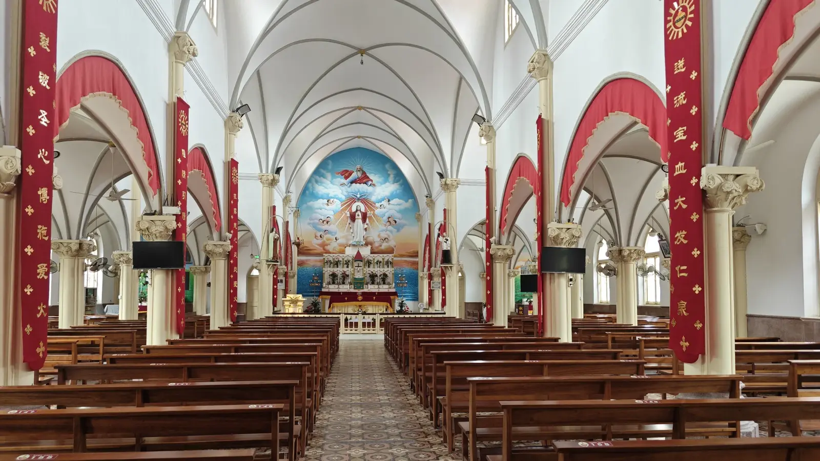 A church interior with cream columns, white arches hung with red drapery, wooden pews and a blue painted apse.