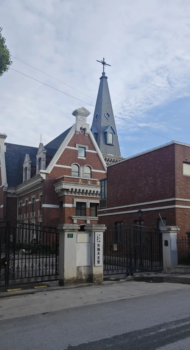 A tall slate spire above a red brick building with white gables, behind a black iron gate and a pale stone gate post.