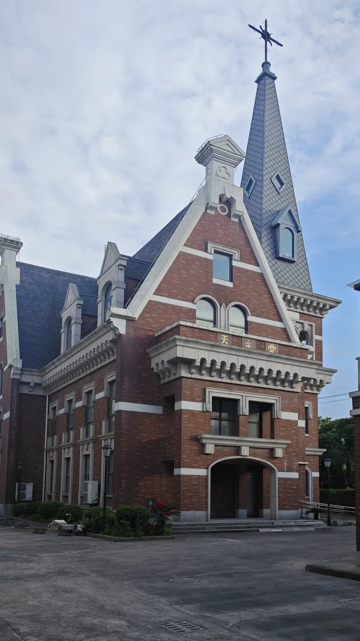 A red brick building with white stepped gables, dormer windows and a tall slate spire topped with a cross, against a blue sky.