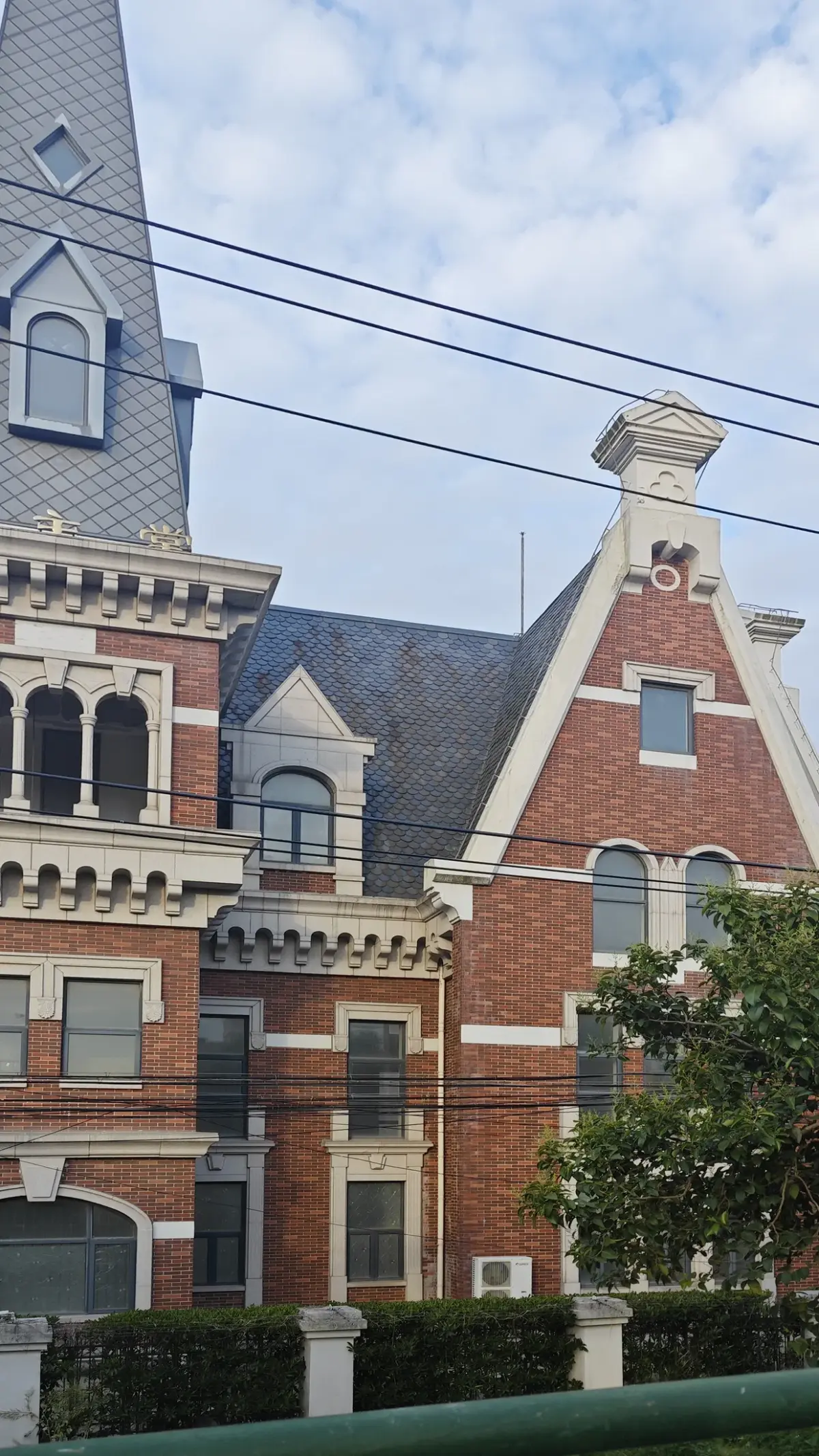A red brick tower with white arches, a small arcade and a slate-clad spire behind a tree and power lines.