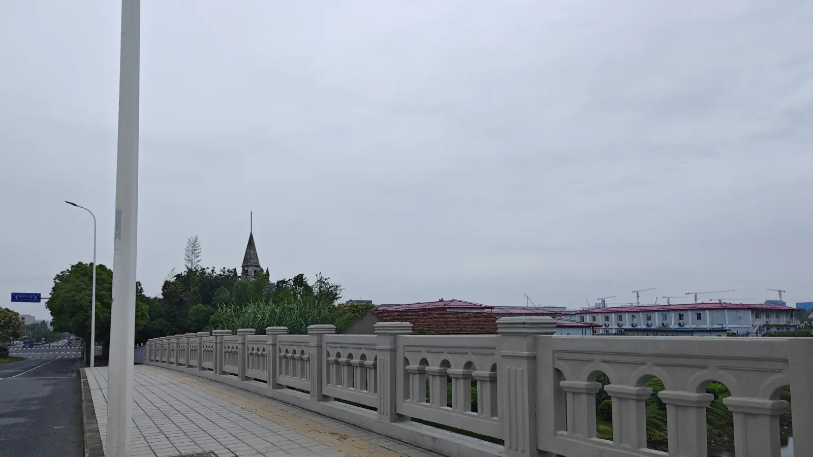A distant church spire above trees beyond a pale stone bridge balustrade and an empty road.