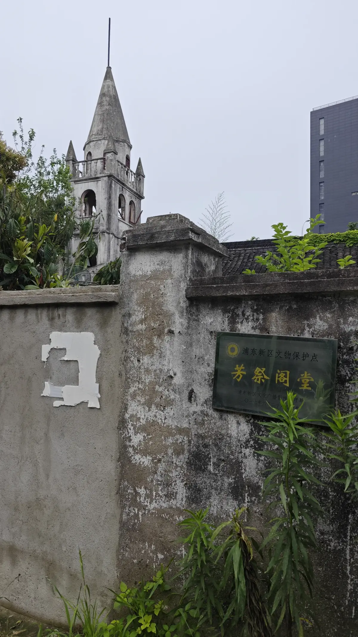 A grey stone steeple with a louvred belfry and cross behind a crumbling wall bearing a green sign.
