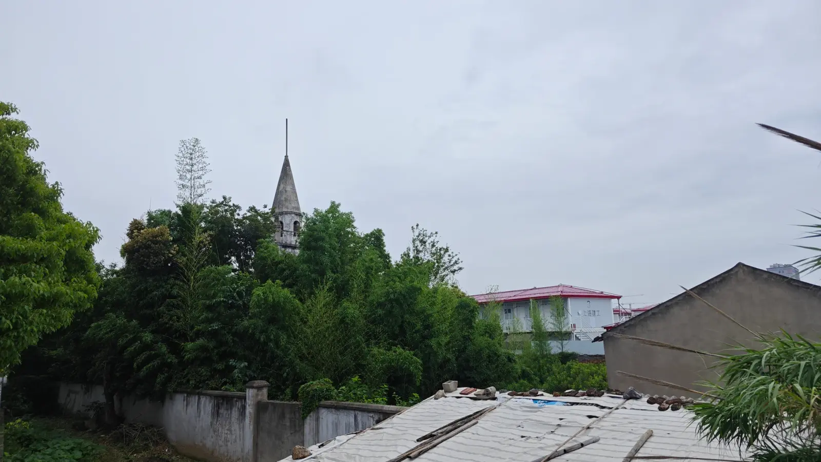 A slender grey spire rising from dense bamboo and trees, with low roofs and a wall in the foreground.
