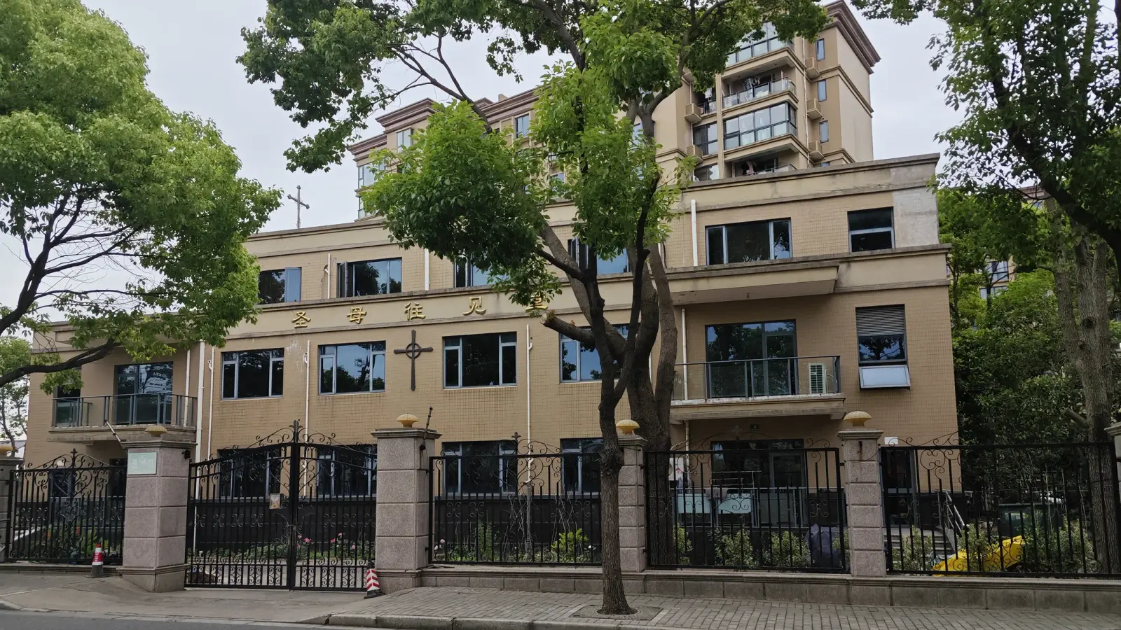 A cream-coloured building of three storeys with gold characters and crosses on its upper wall, behind iron railings and a tree.