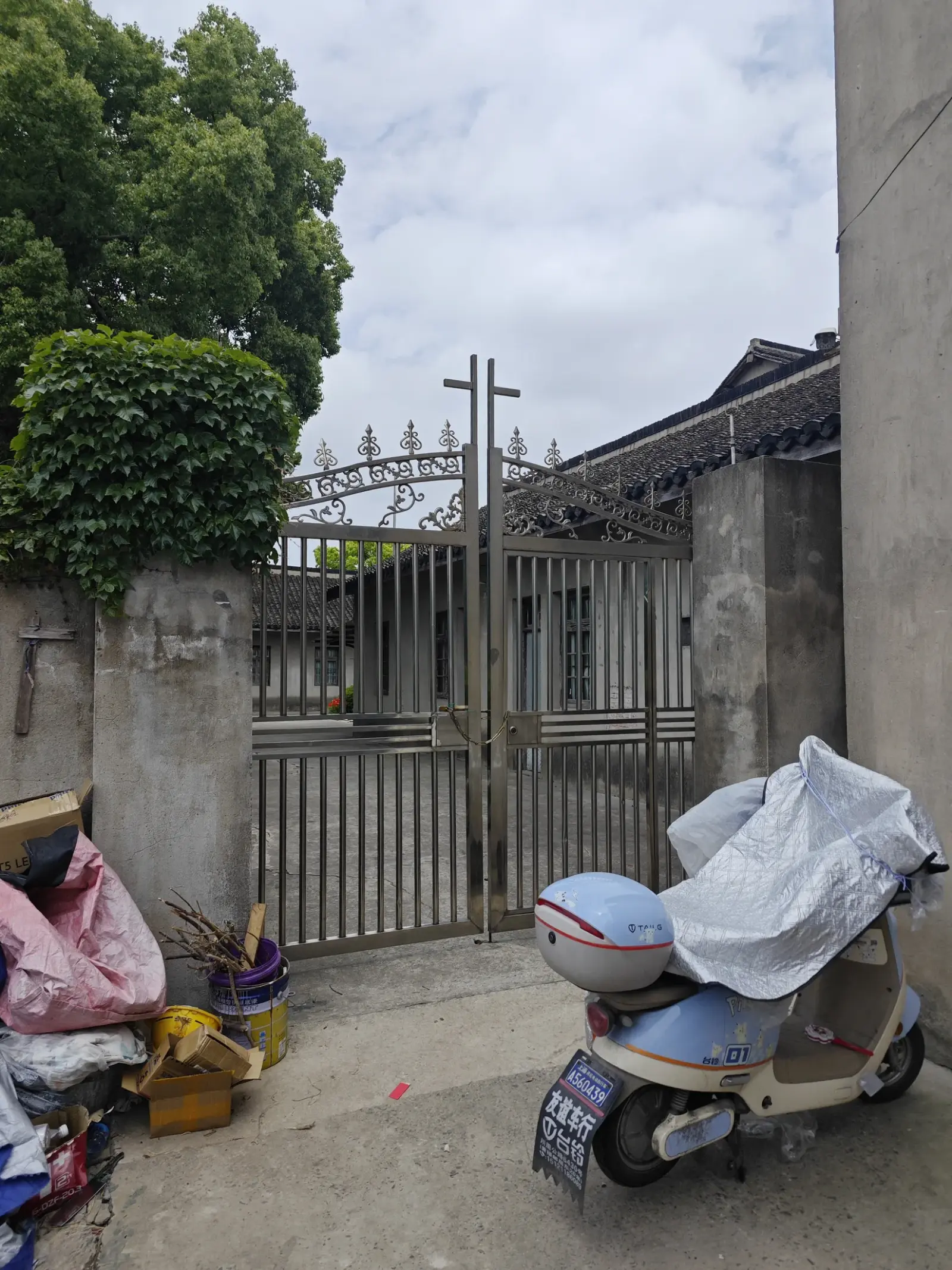 A tall metal cross above a closed iron gate between concrete gate posts, with a scooter and bundled belongings in front.