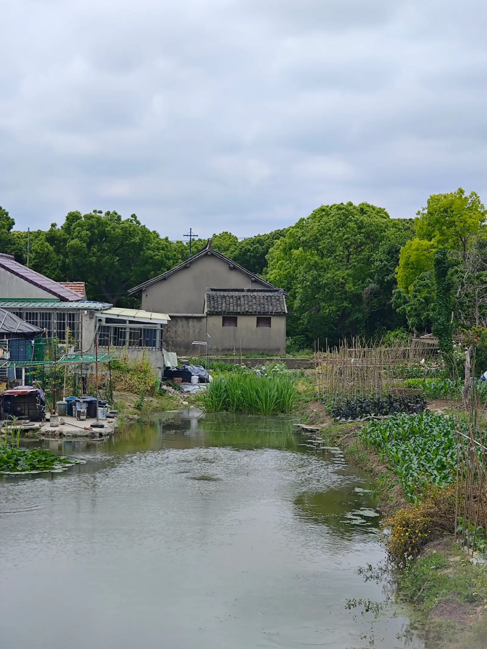 A grey gabled building with a cross beside a canal, with allotments, trees and other village houses around it.
