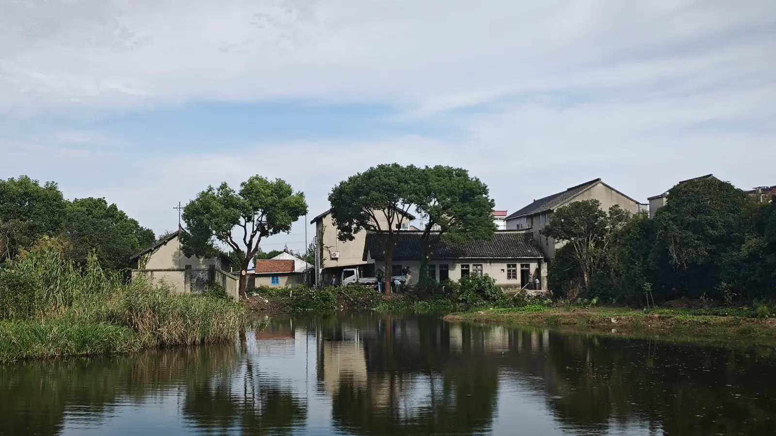 Low grey-walled buildings with tiled roofs and tall trees reflected in a pond under a cloudy sky.