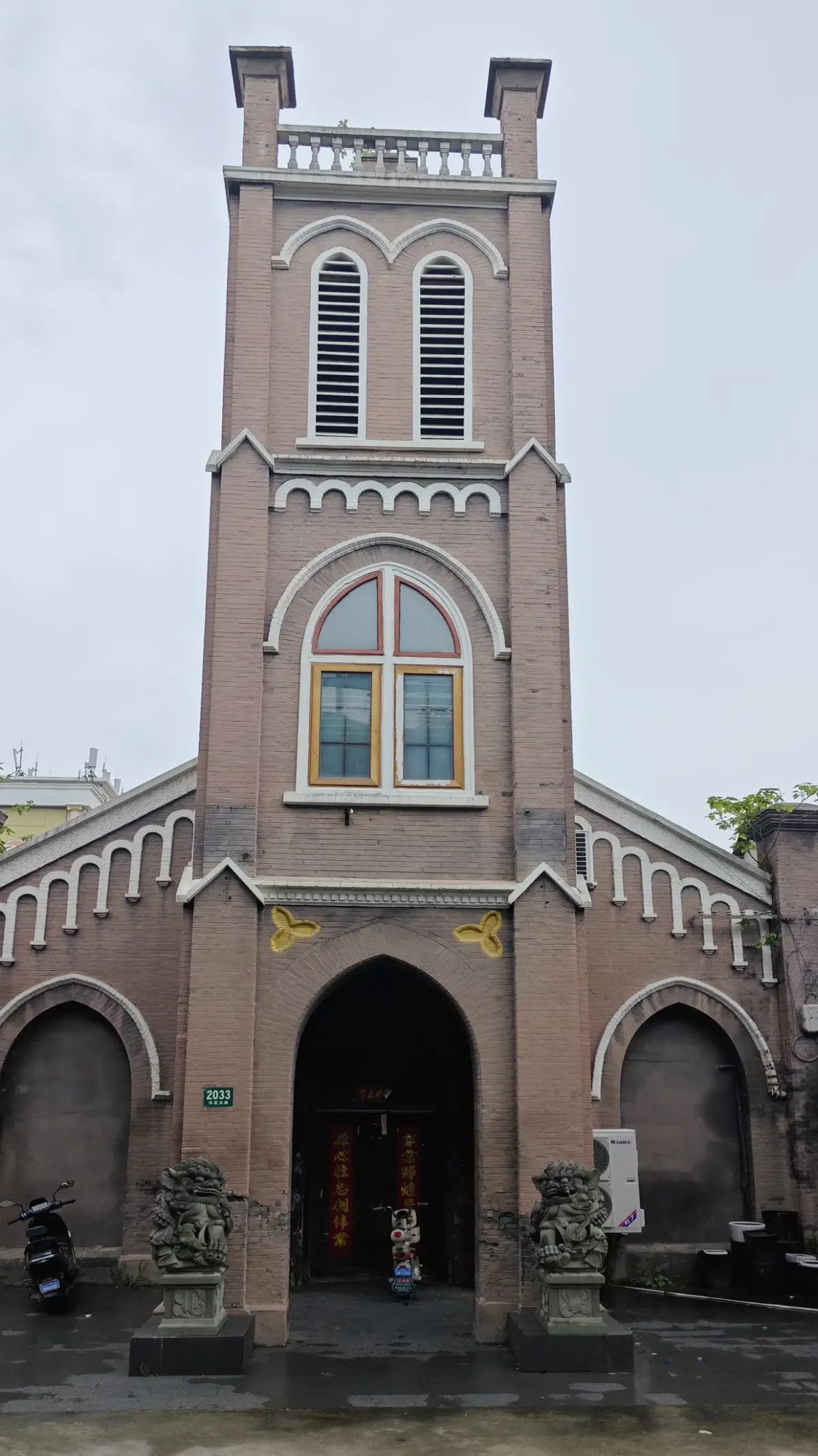 A tall brick church tower with white-trimmed arched windows and a pointed-arch doorway, with two small stone lions in front.
