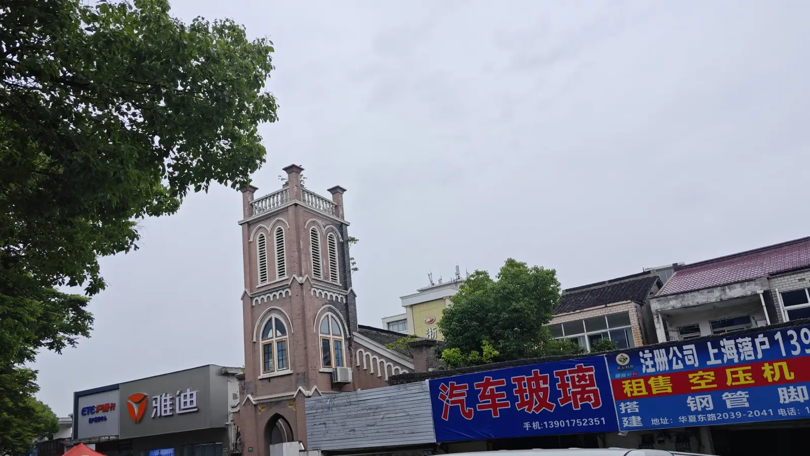 A brick church tower with arched windows rising behind a tree and above colourful shop signs in a street.