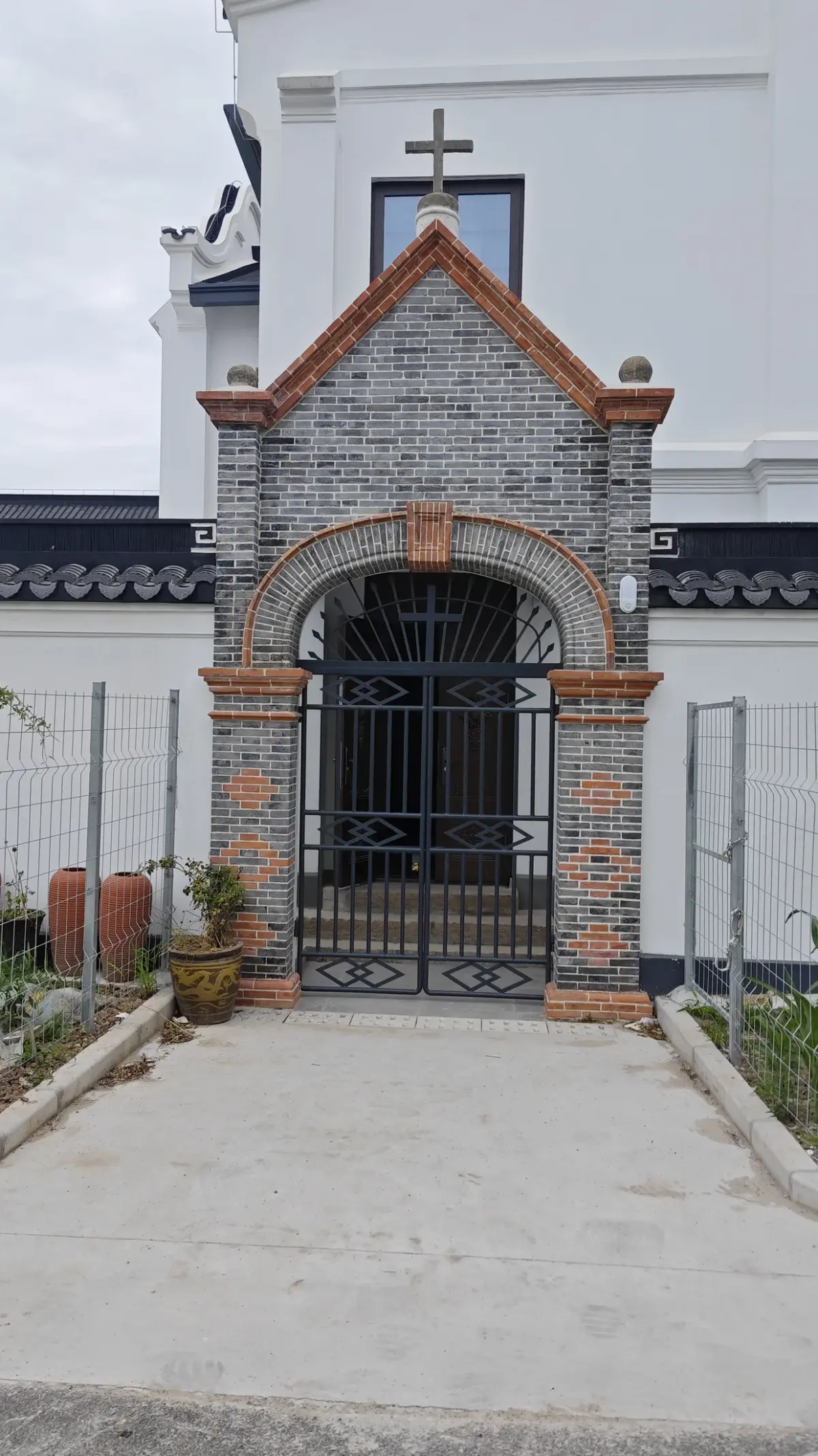 A grey brick archway edged in orange brick with an iron gate, set in front of a white church wall topped by a cross.