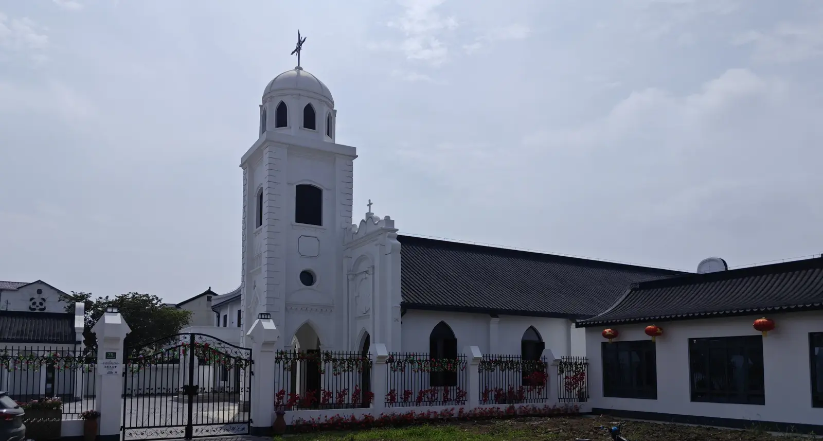 A tall white bell tower with a small dome and cross at the left of a long white building with a dark tiled roof, behind an iron gate and fence.