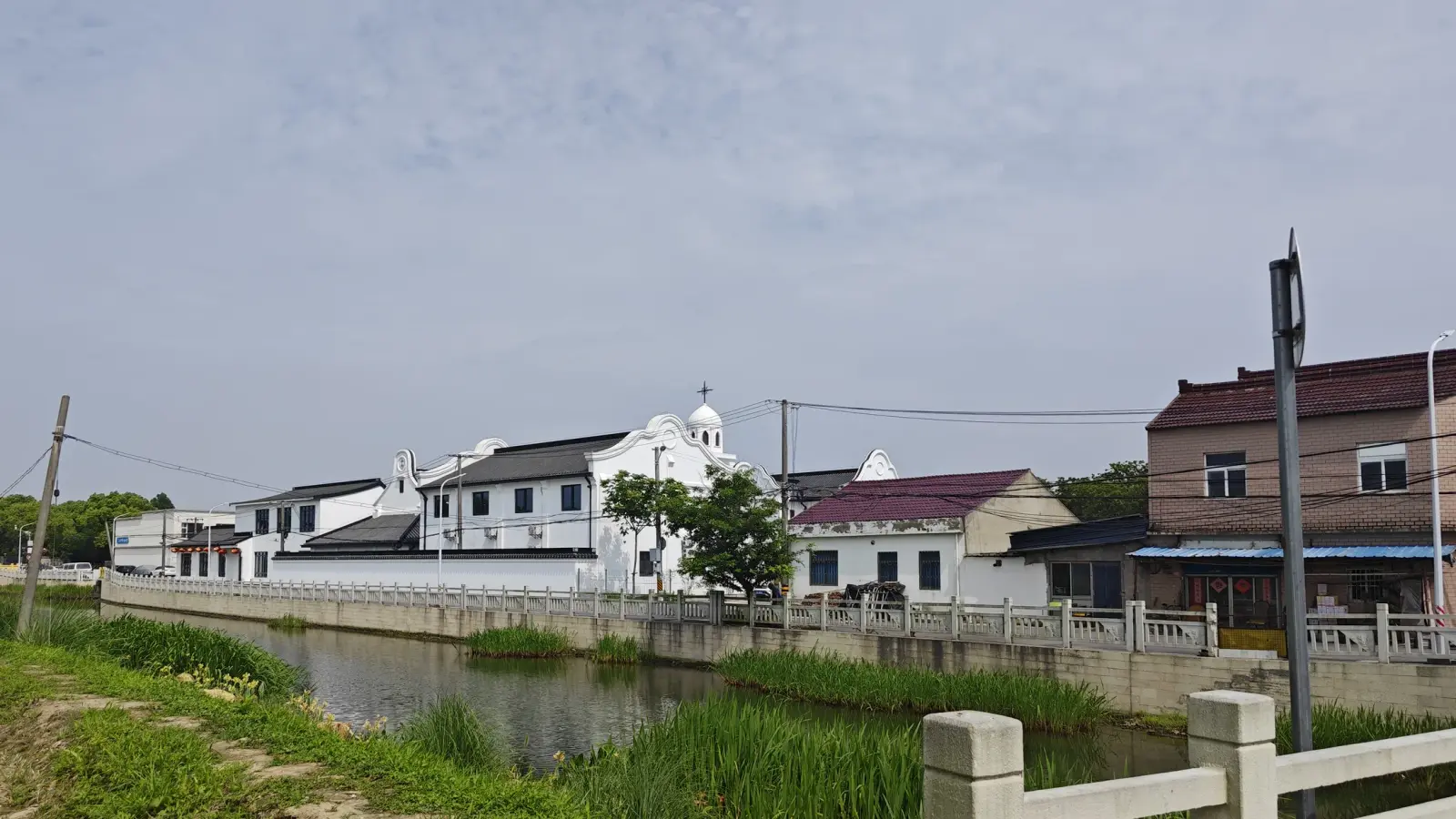 White buildings with black tiled roofs and a small domed tower beside a canal with a stone railing and reeds.