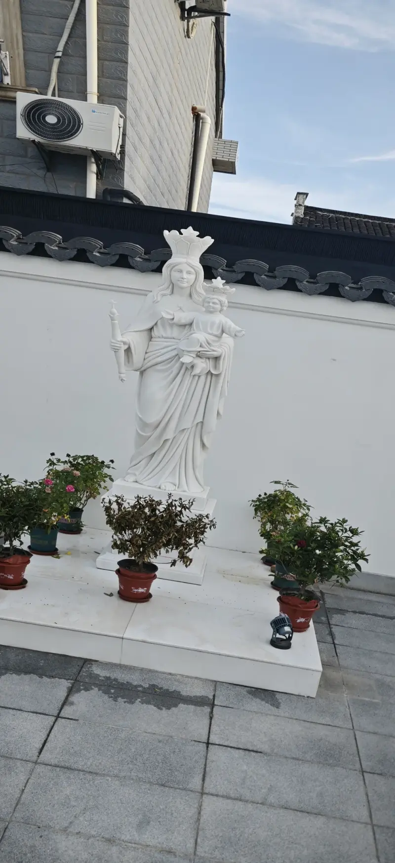 A crowned white statue of a woman holding a child, on a white base with potted plants, against a white wall with tiled coping.