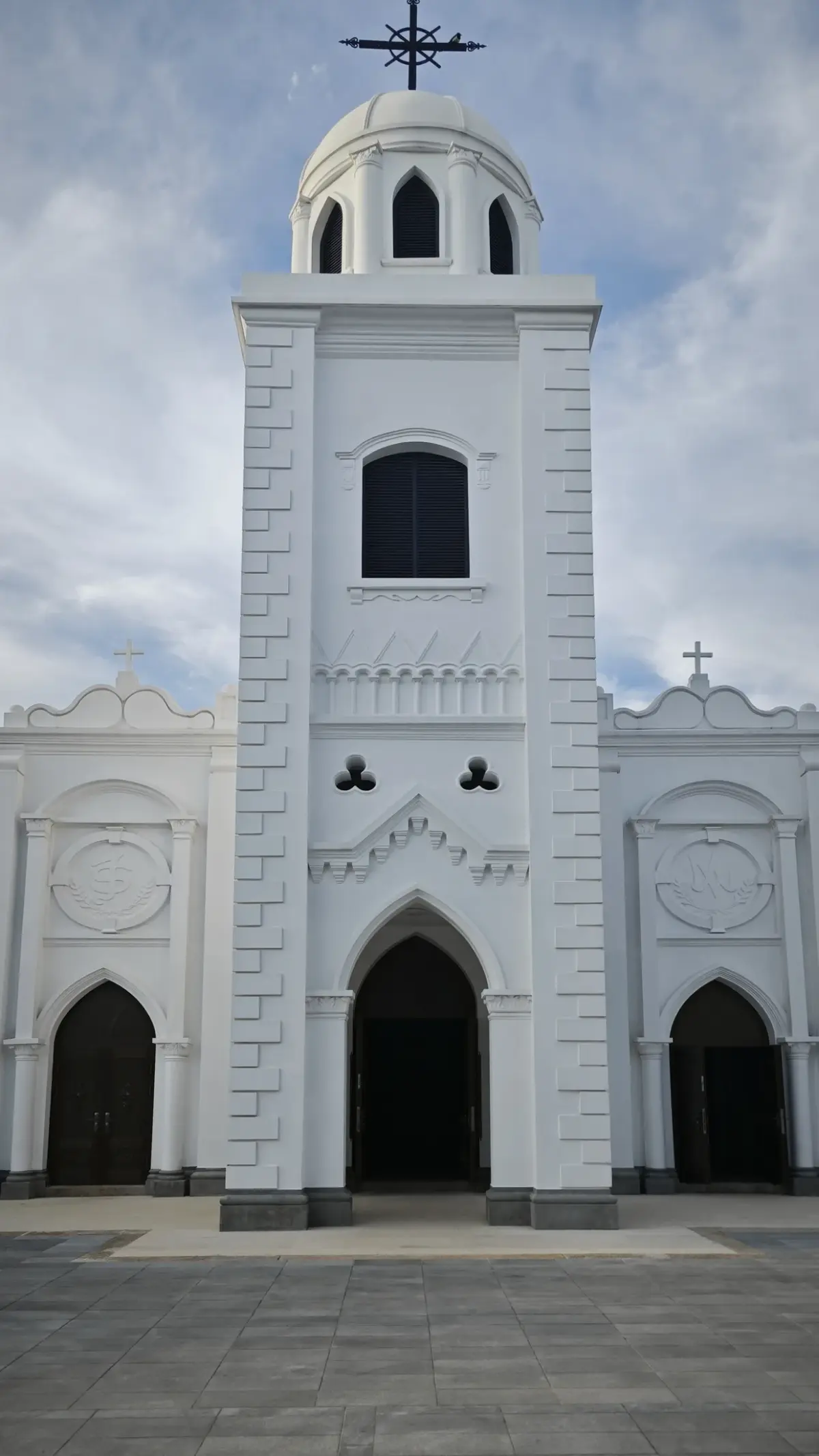 A white church front with a tall central tower topped by a dome and cross, and two pointed-arch doors beneath decorated gables.