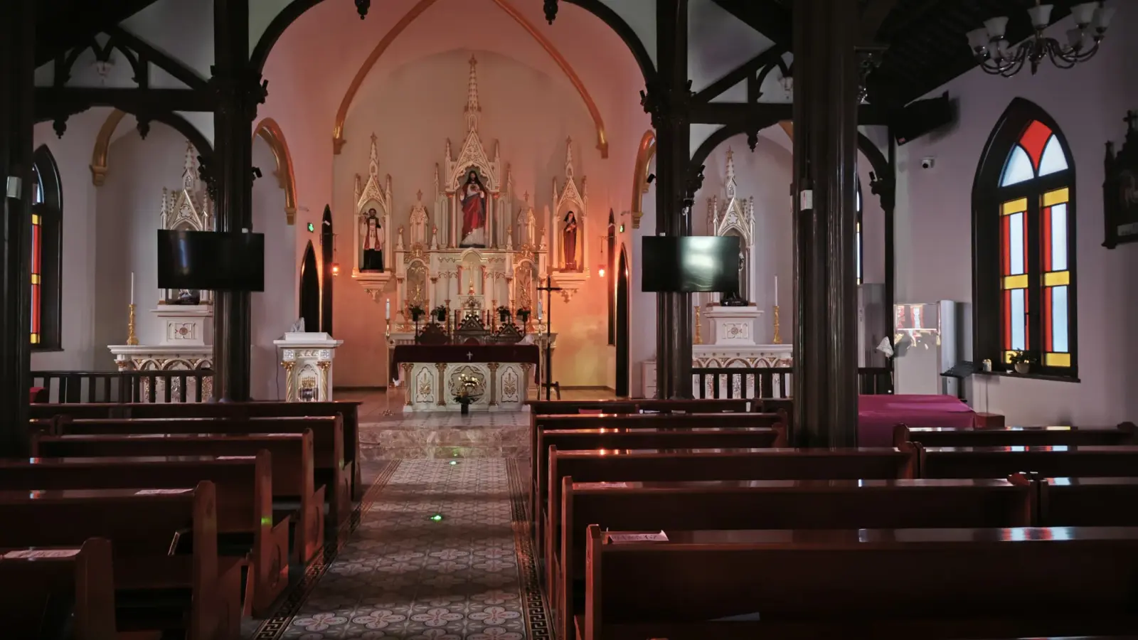 A pale Gothic altarpiece with a red-robed statue in the centre, flanked by smaller altars and stained glass windows in red and yellow.
