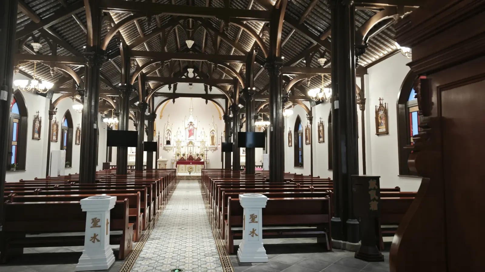 A church interior with dark timber arches and trusses lit from below, wooden pews and a tiled aisle leading to a white altar.