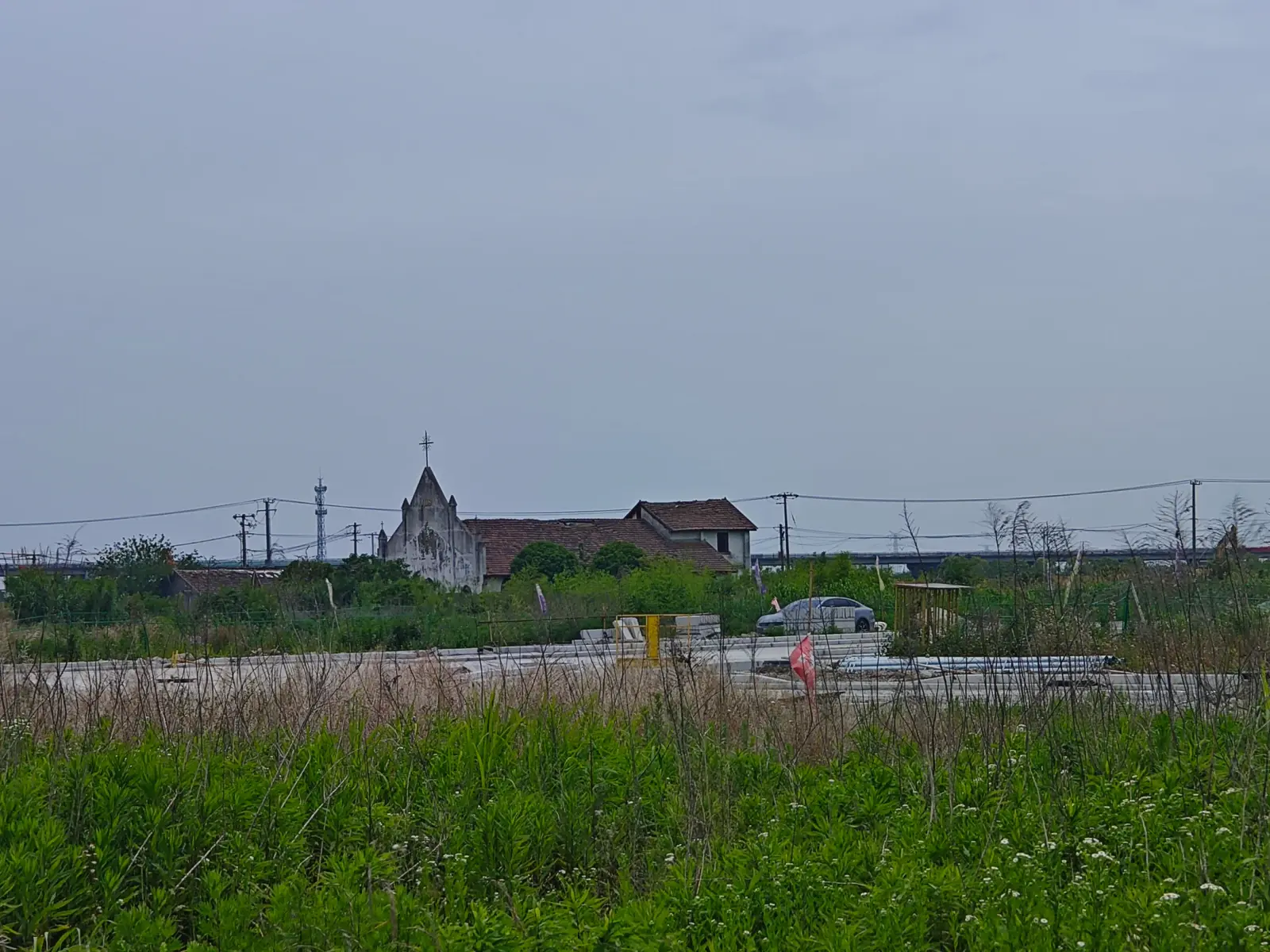 A small grey pointed church gable with a cross beside red-tiled roofs, seen across a cleared field under a heavy grey sky.