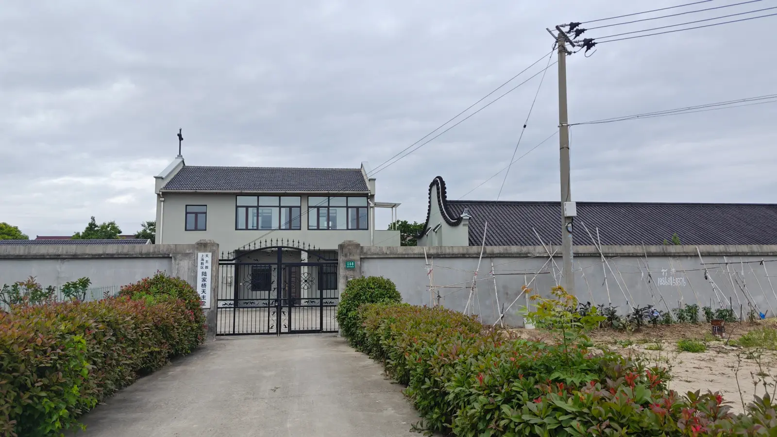 A two-storey grey building with a cross on its dark tiled roof behind an iron gate and a long grey wall, at the end of a tree-lined concrete path.