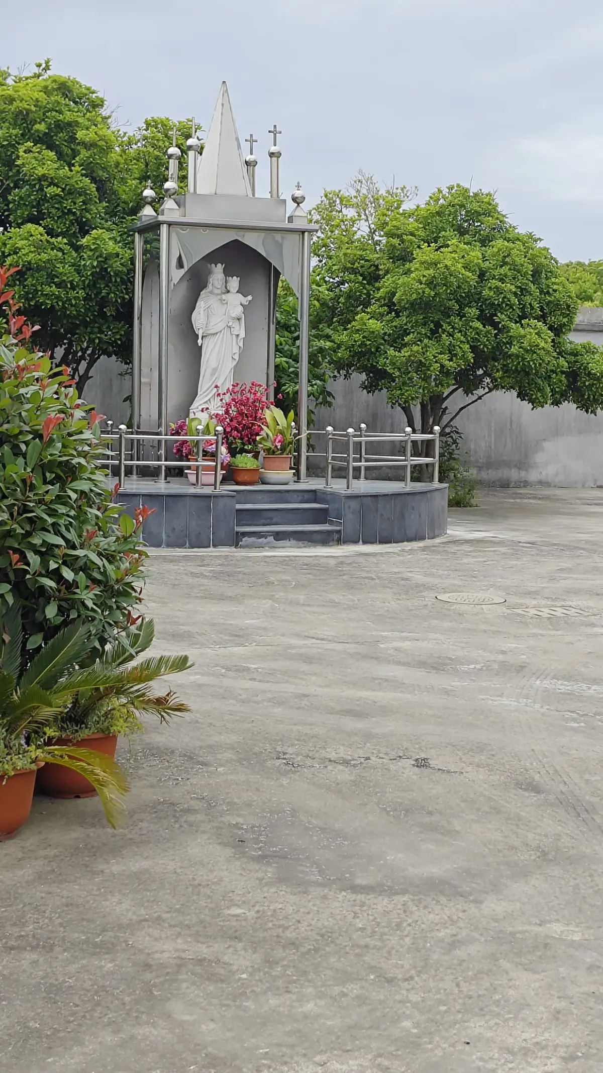 A pale stone shrine with a pointed spire housing a white statue, with red flowers on its base, in an open concrete yard.