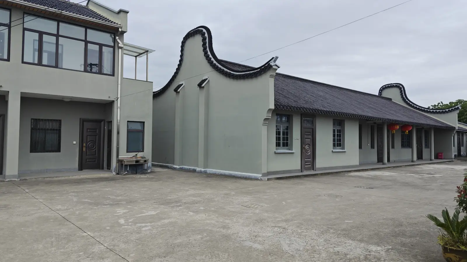 A long grey-green single-storey building with black tiled roof and scrolled gable ends beside a two-storey block and a concrete yard.