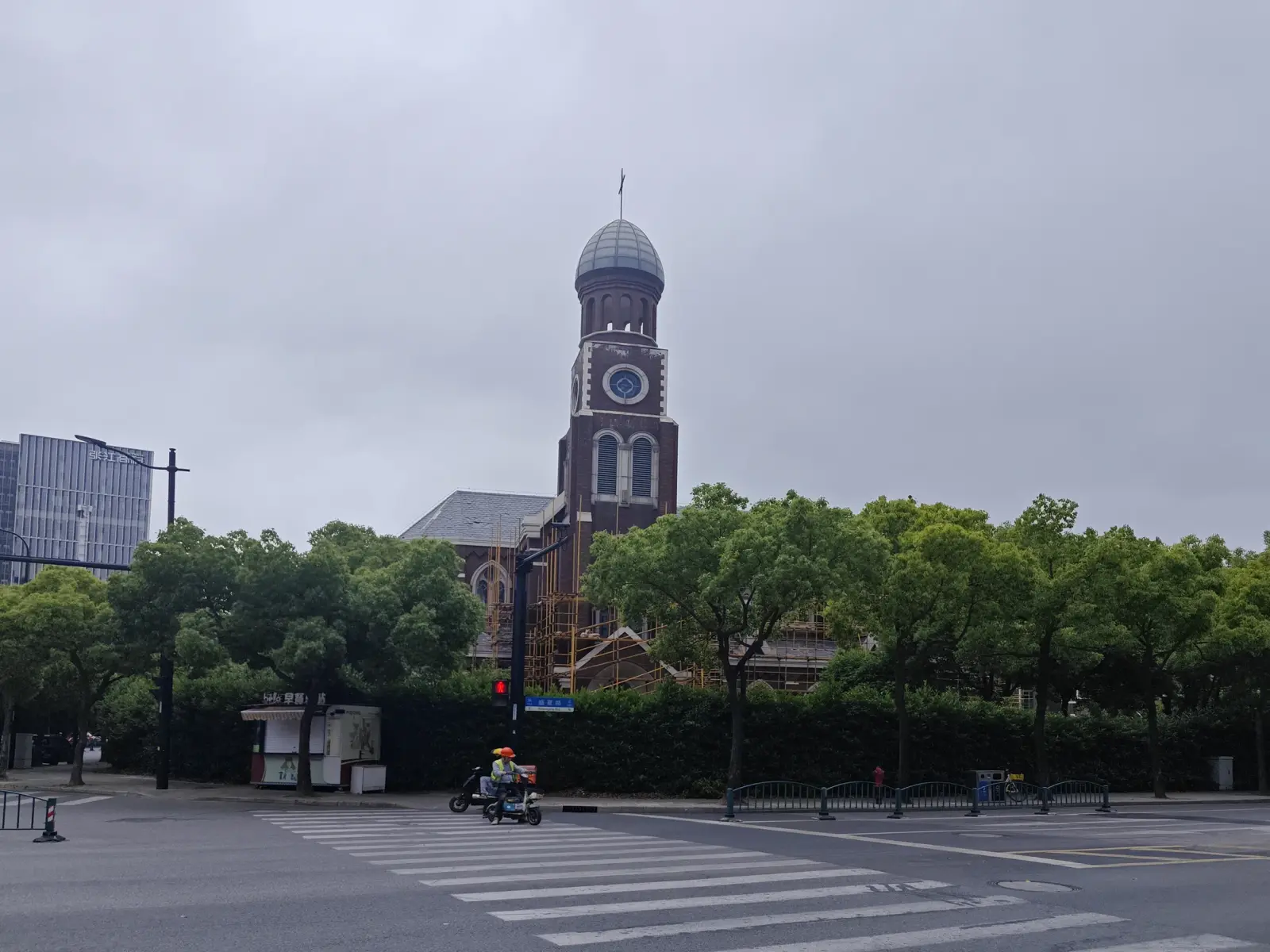 A brick church tower with a grey dome, cross and clock above a hedge and trees, beyond a pedestrian crossing and modern buildings.