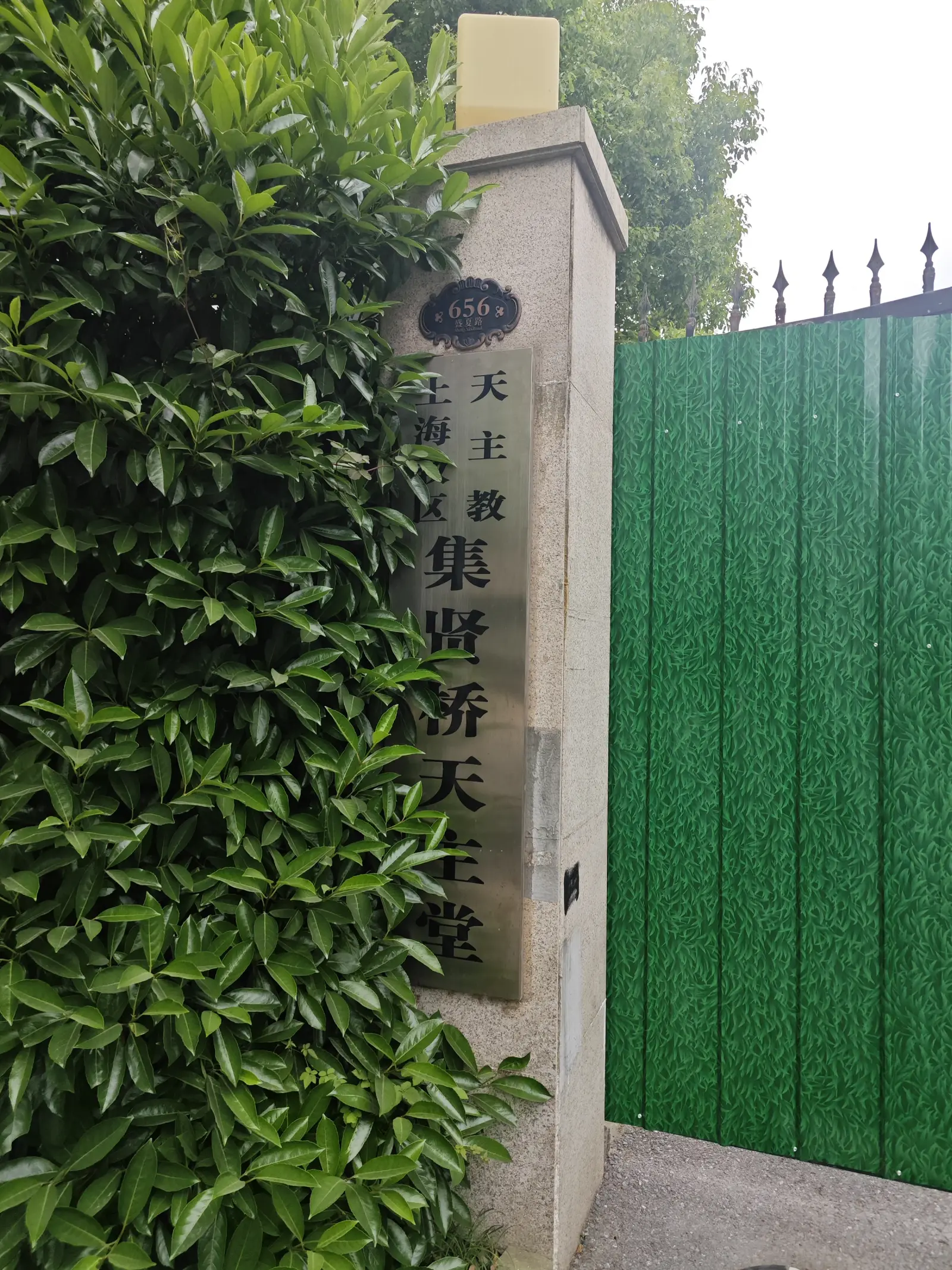 A pale stone gate pillar with a vertical black name plate in Chinese, with green foliage on the left and a green metal gate on the right.
