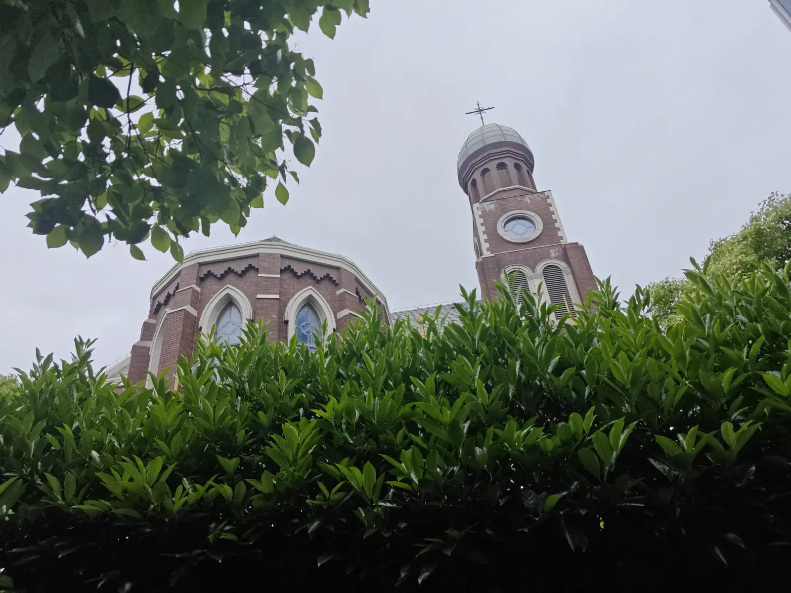 A brick church tower with a clock face and a grey domed top, and a pointed-arched window gable, above a green hedge.
