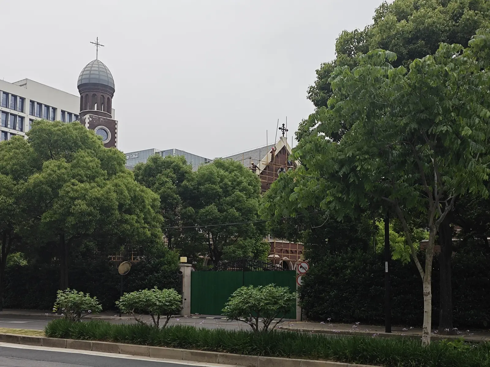 A brick church with a grey domed tower, a smaller gable with a cross and a green steel gate, behind street trees and a hedge.