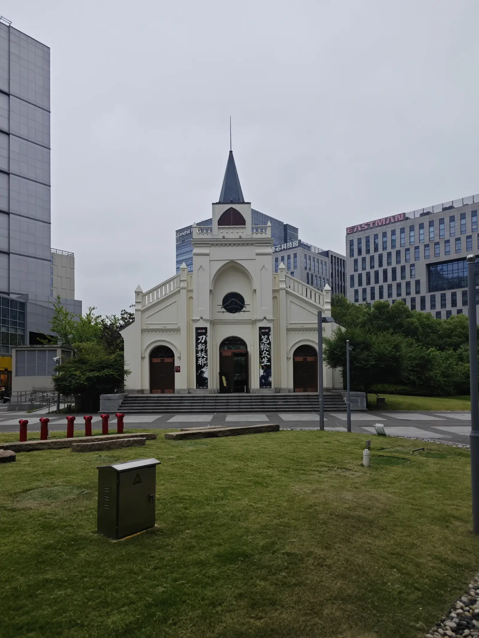 A small cream church with a pointed spire and arched doorways on a lawn, surrounded by tall modern office buildings.