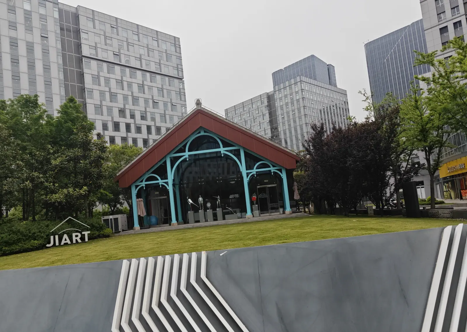 A pavilion with a red pitched roof and turquoise iron arches on a lawn, beneath tall glass office buildings.