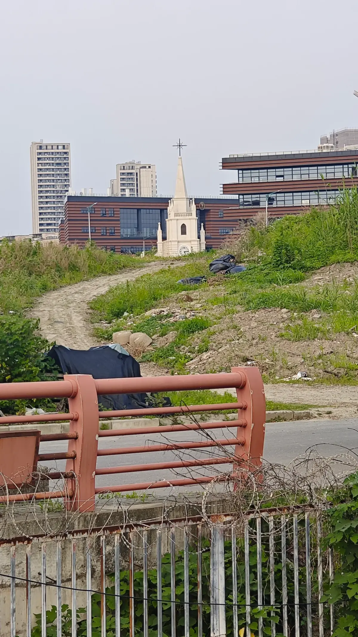 A small white church with a pointed spire and cross beyond a dirt track and a red railing, with modern buildings behind.