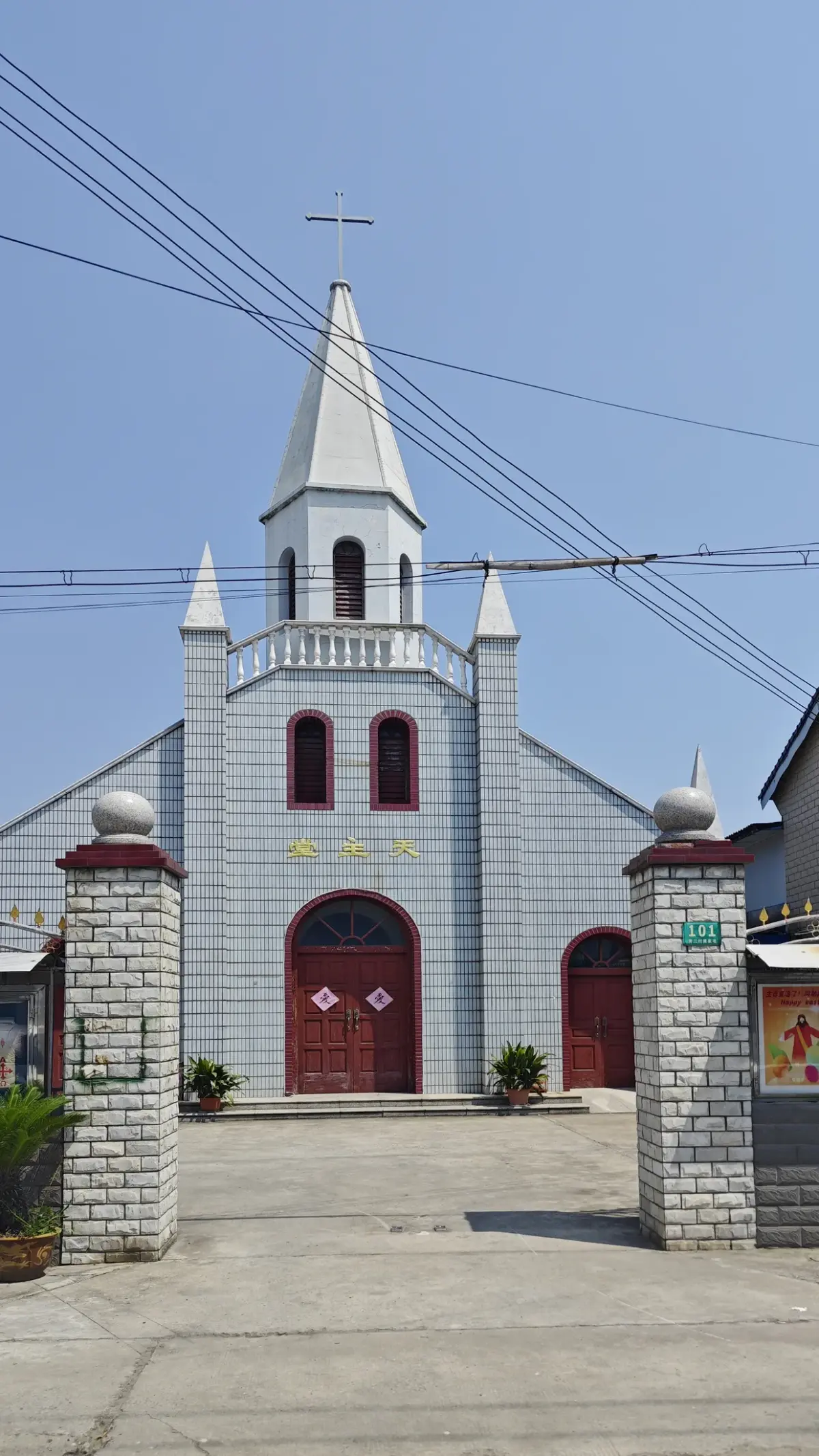 A grey church front with a tall white spire, two narrow red windows and red arched doors between brick gate piers, with overhead wires.