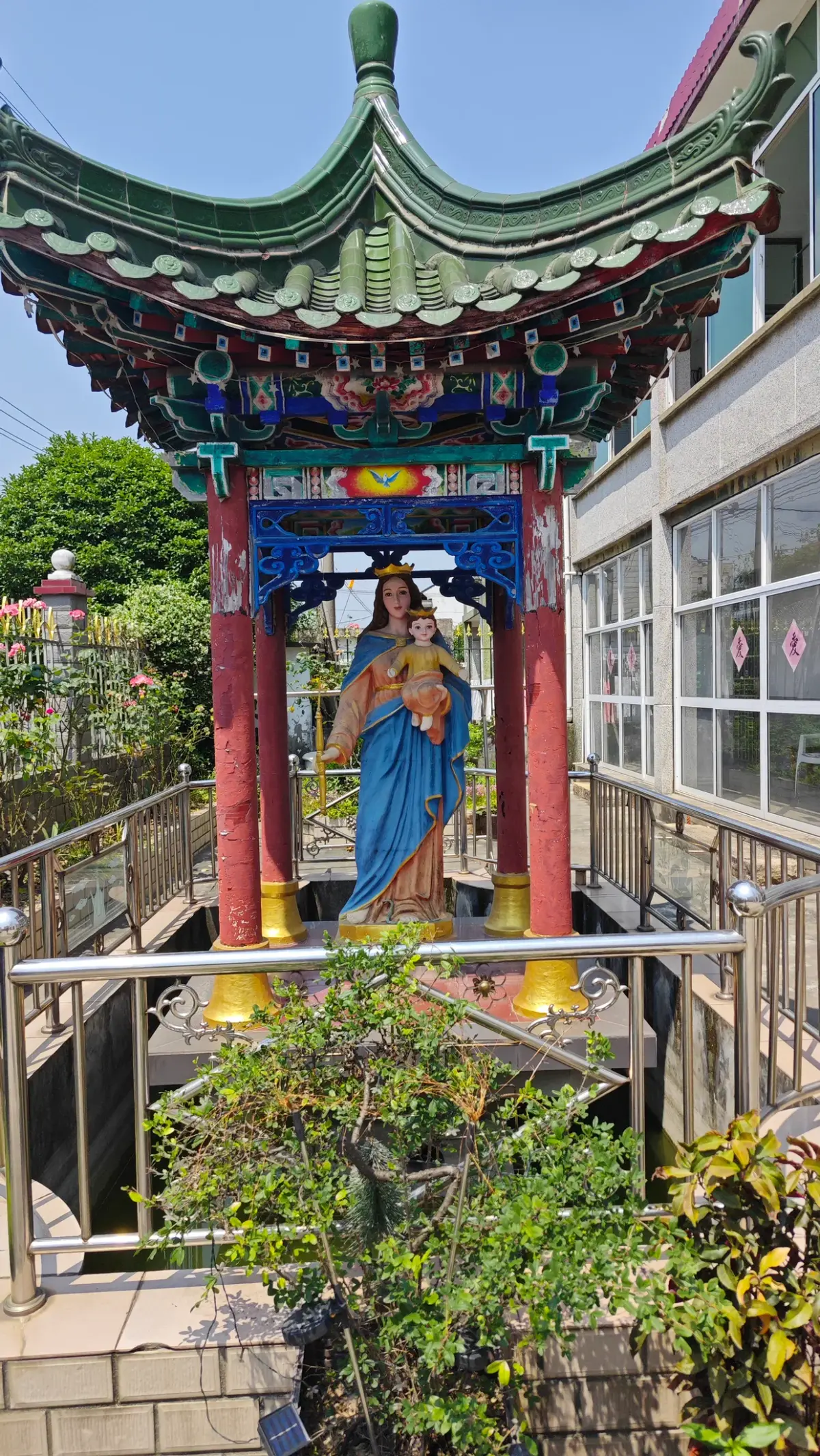 A colourfully painted statue of a woman in blue holding a child, under a pavilion roof with red pillars and green tiles.