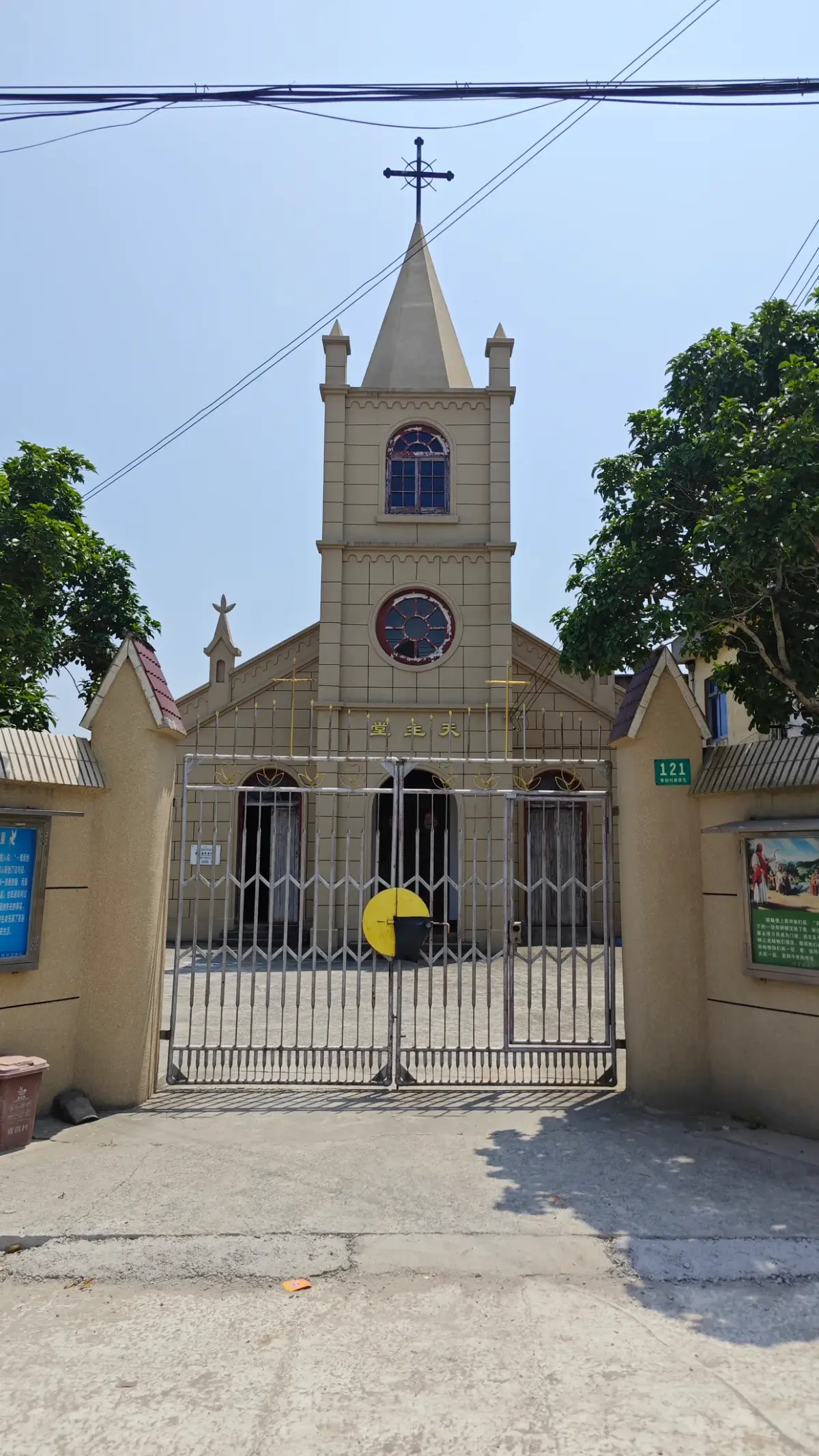 A cream church with a pointed steeple and cross behind a closed iron gate between low gate walls, under a blue sky.
