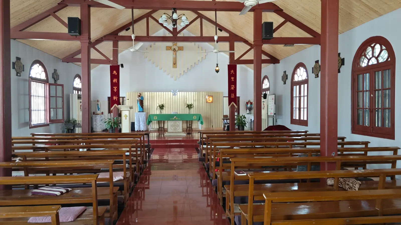 A church interior with red-brown timber columns and roof trusses, wooden benches, a crucifix and white walls with arched windows.