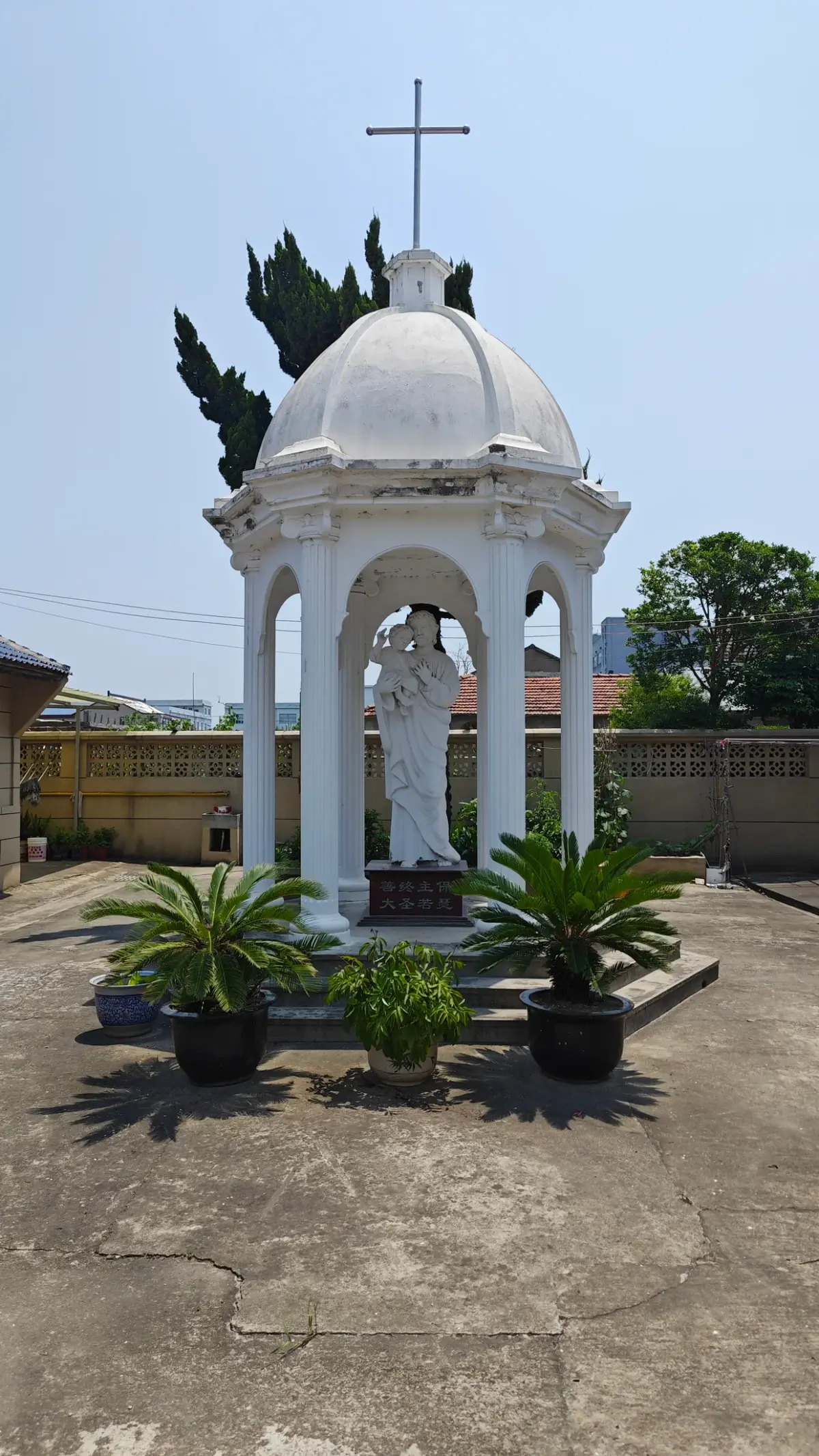 A small white domed open pavilion with a cross on top housing a white statue, with potted palms around it.