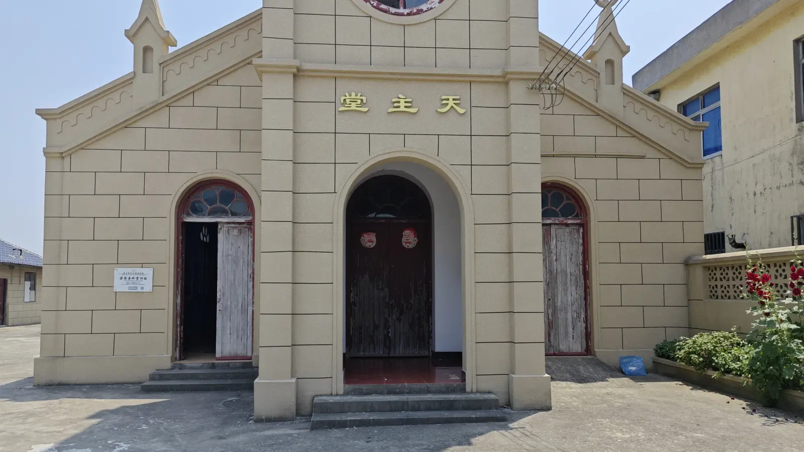 A cream-coloured church front with a central tower, three arched doorways and gold Chinese characters on the tower, in strong sunlight.