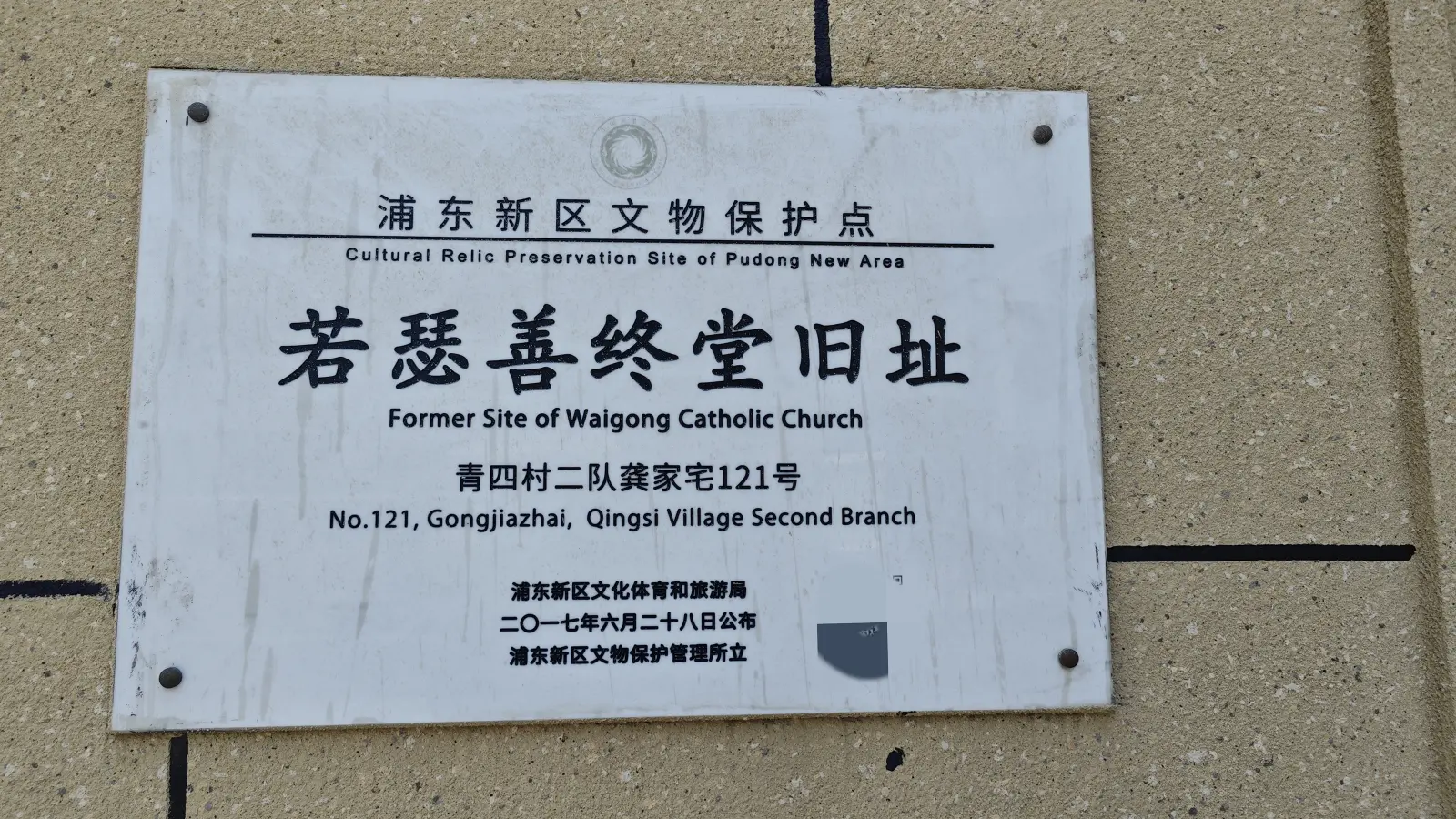 A white plaque with Chinese and English text on a sand-coloured wall, naming the former church site and its address.