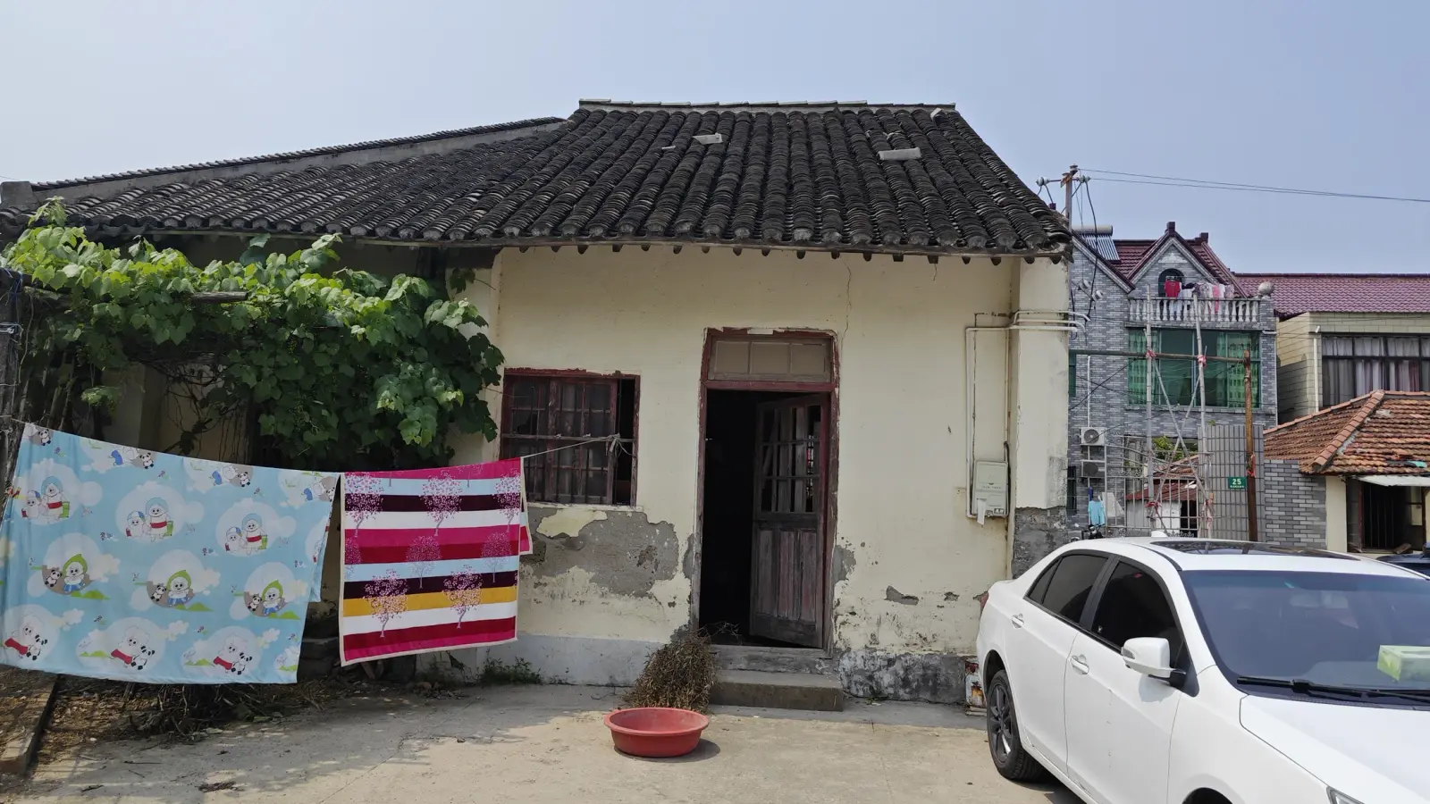 A cream building with flaking plaster, a dark wooden doorway and a grey tile roof, with washing hung out and a white car at right.
