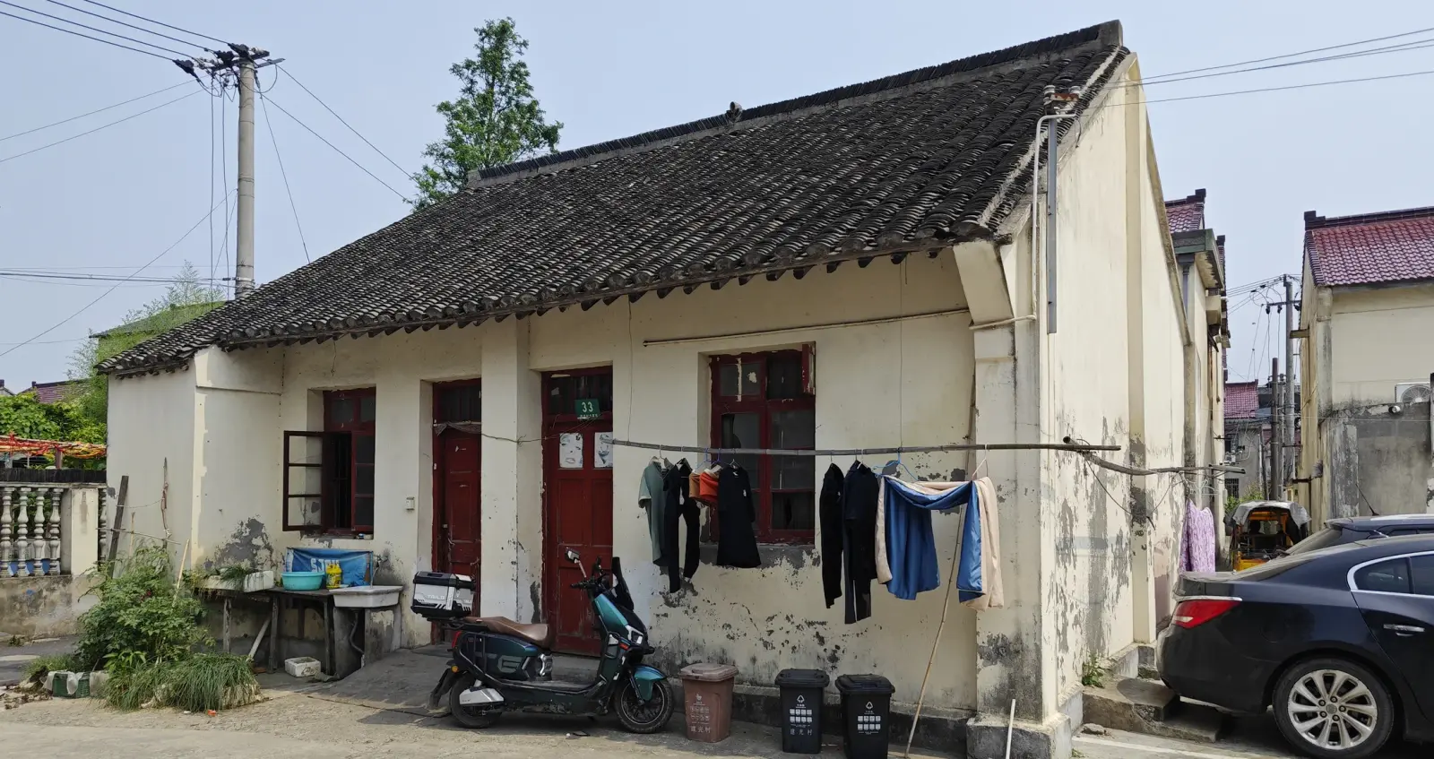 A single-storey cream building with a dark grey tile roof, red doors and laundry hanging outside, beside a parked car.