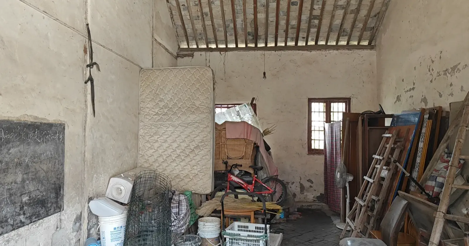 A whitewashed room with a timber and tile ceiling, a blackboard on one wall and furniture and tools stacked inside.