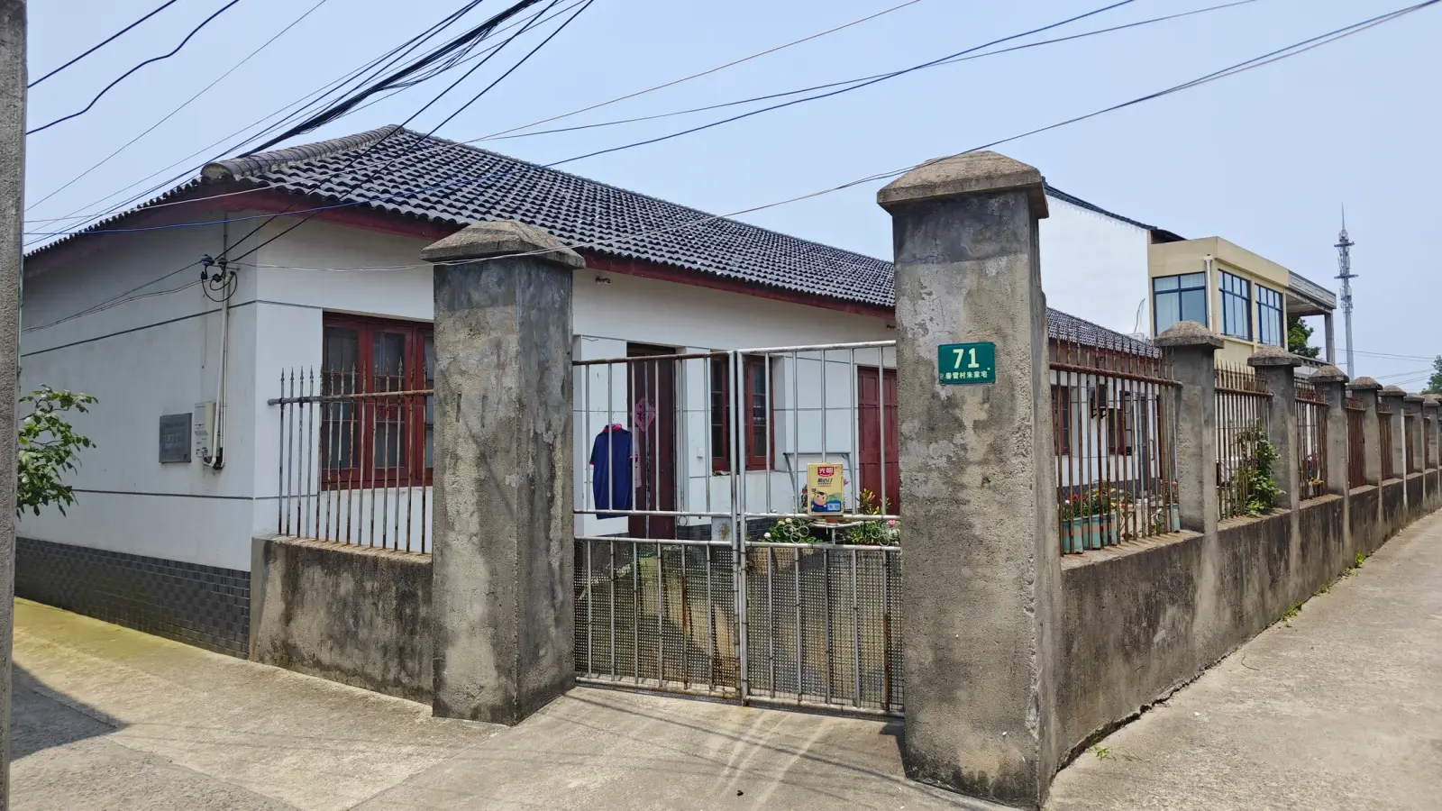 A whitewashed single-storey building with a grey tiled roof and red doors behind stone gate posts and an iron fence.